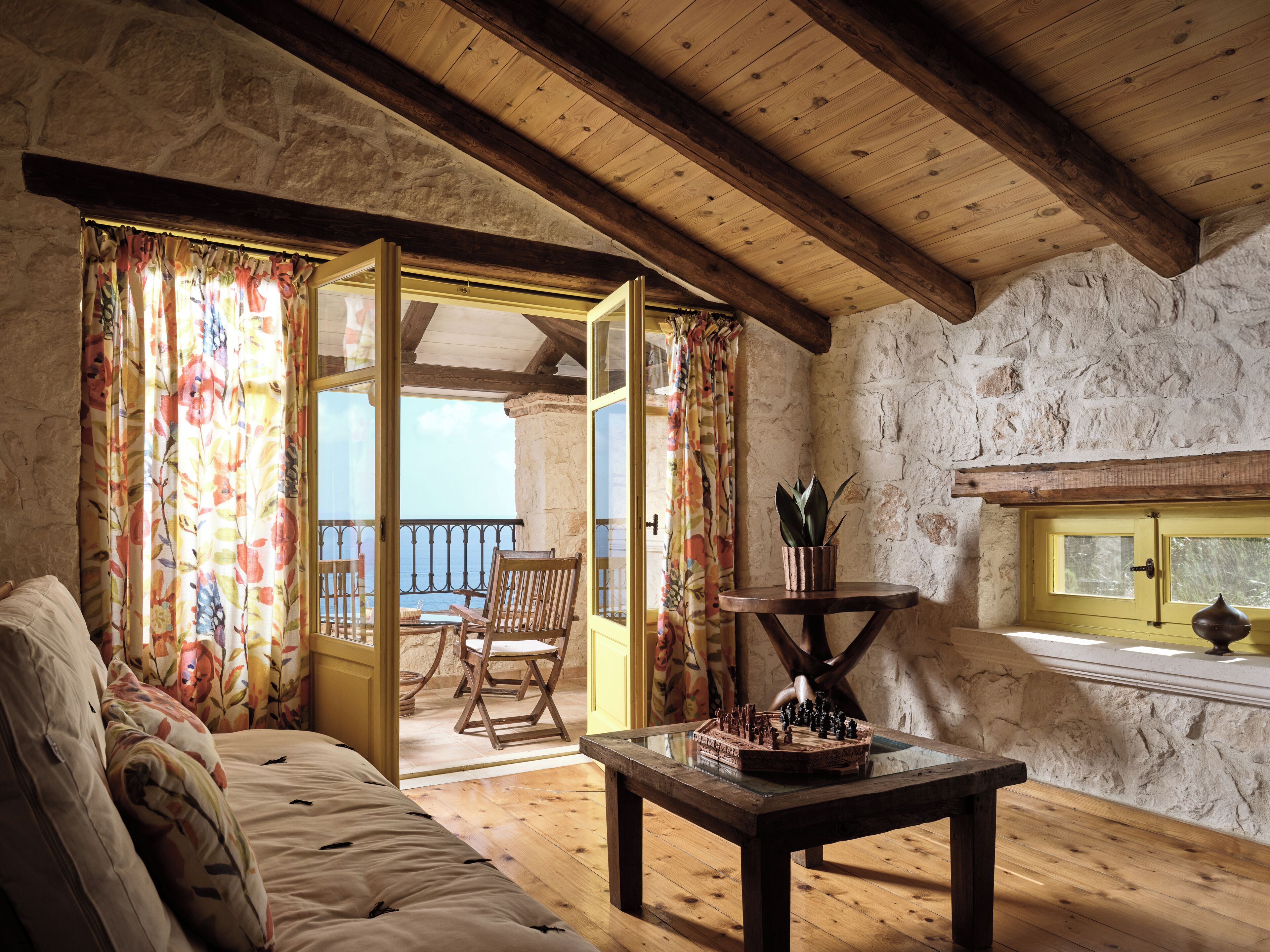 A cozy room with a wooden ceiling, stone walls, a wooden sofa, a table with a chessboard, and a view of the sea through an open door.