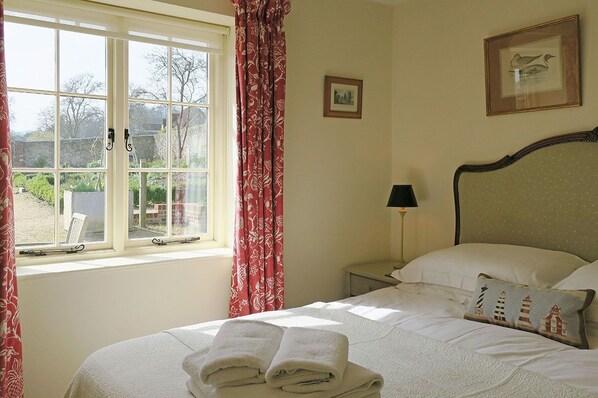 A neatly made bed with folded towels, a bedside table with a lamp, and a window with curtains. - Folly Cottage (Eastbourne)
