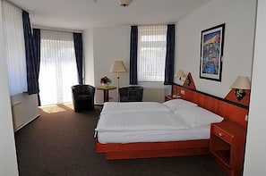 Quarto de hotel com cama, duas cadeiras, escrivaninha, luminárias e um quadro na parede.