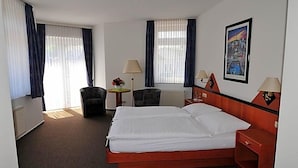 A hotel room with a bed, two chairs, a desk, lamps, and a painting on the wall.