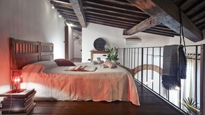 Ein Schlafzimmer mit Holzboden, einem Bett mit rosa Bettdecke, einem Nachttisch mit Büchern und einem Balkon mit Geländer.
