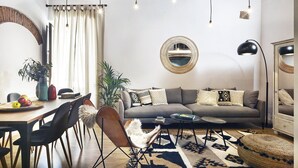 Ein modernes Wohnzimmer mit einer Couch, einem Couchtisch und Sesseln. Ein Esstisch mit Stühlen und einer Pflanze steht ebenfalls im Raum. Ein großes Spiegel und Pendelleuchten vervollständigen das Bild.