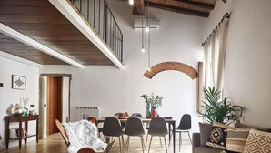 Ein geräumiger Wohnbereich mit hoher Decke, Holzbalken, einem Esstisch mit Stühlen, einer Couch mit gemusterten Kissen und einer Treppe, die zu einer oberen Ebene führt.