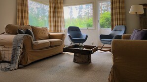 A cozy living room with a sofa, two armchairs, a coffee table, and a mounted deer head. Large windows offer a view of trees. - Vildmarks Lodge (Ytterhogdal)