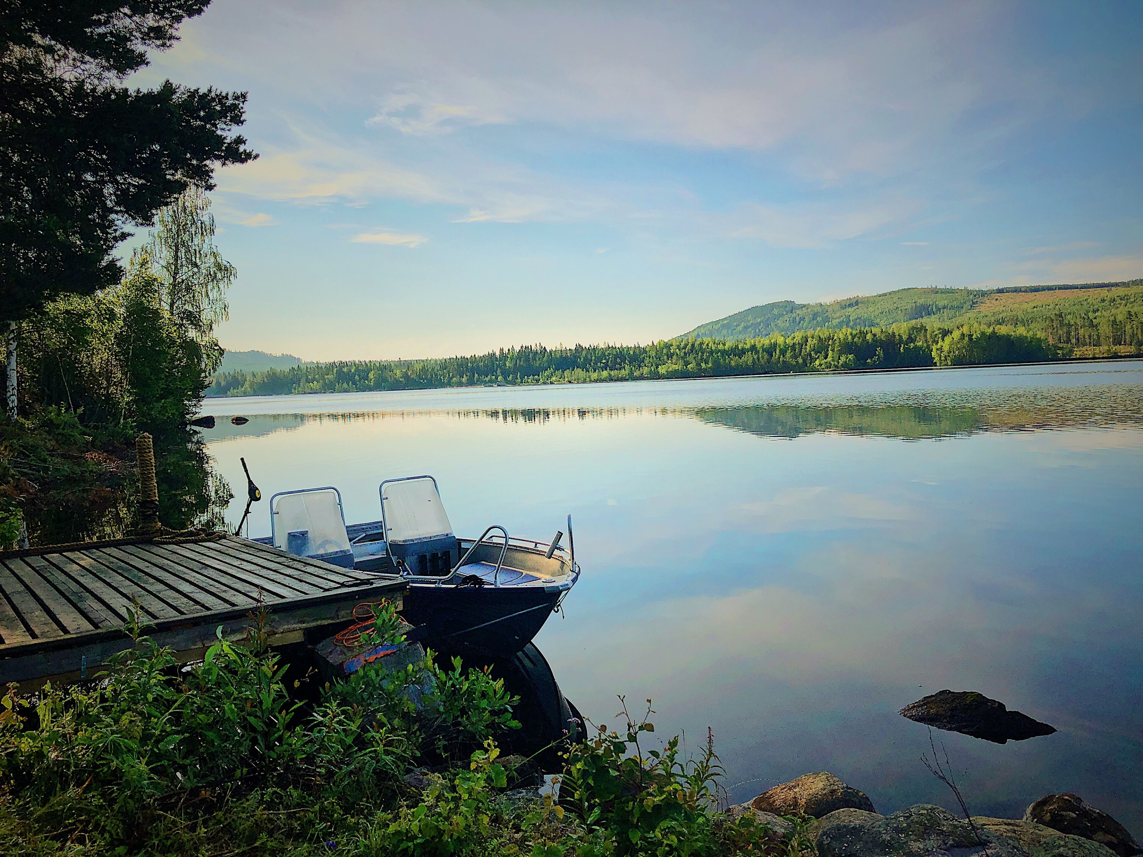 En stilla sjö med en brygga och stolar, omgiven av träd och kullar.