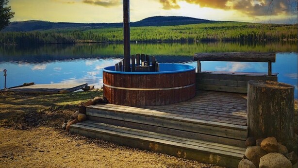 A wooden hot tub on a dock by a calm lake with a forested hill in the background. - Vildmarks Lodge (Ytterhogdal)