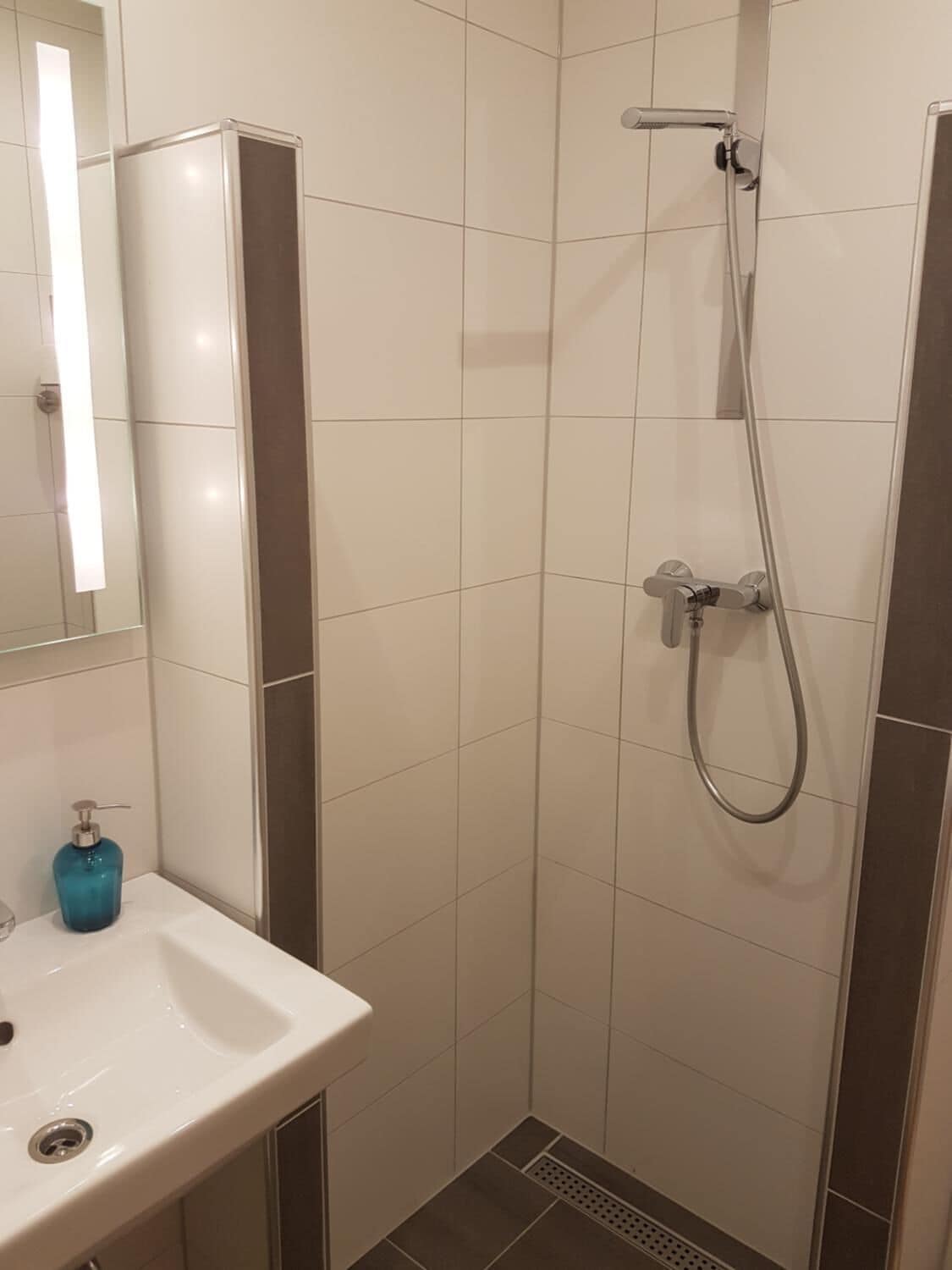 Un baño con un lavabo blanco, un dispensador de jabón azul, un espejo y una ducha con cabezal de ducha manual.
