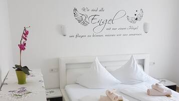 Habitació d'hotel amb un llit blanc, dos coixins, una taula de nit amb un vas verd i una orquídia rosa, i una samarra mural amb text alemany i cors alats.