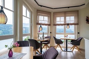 Ein Essbereich mit Tisch und Stühlen, großen Fenstern mit Vorhängen und Blick ins Freie.