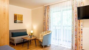 Una habitación con sofá, sillón, una mesita, televisor montado en la pared y ventana con cortinas.