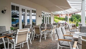 Un espacio de estar al aire libre techado con sillas y mesas, ventanas amplias y vistas del paisaje circundante.