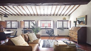 Een gezellige woonkamer met een bruine leren bank, een ronde salontafel en een houten dressoir. Er zijn ramen met radiatoren, een plant en een spiegel aan de muur.