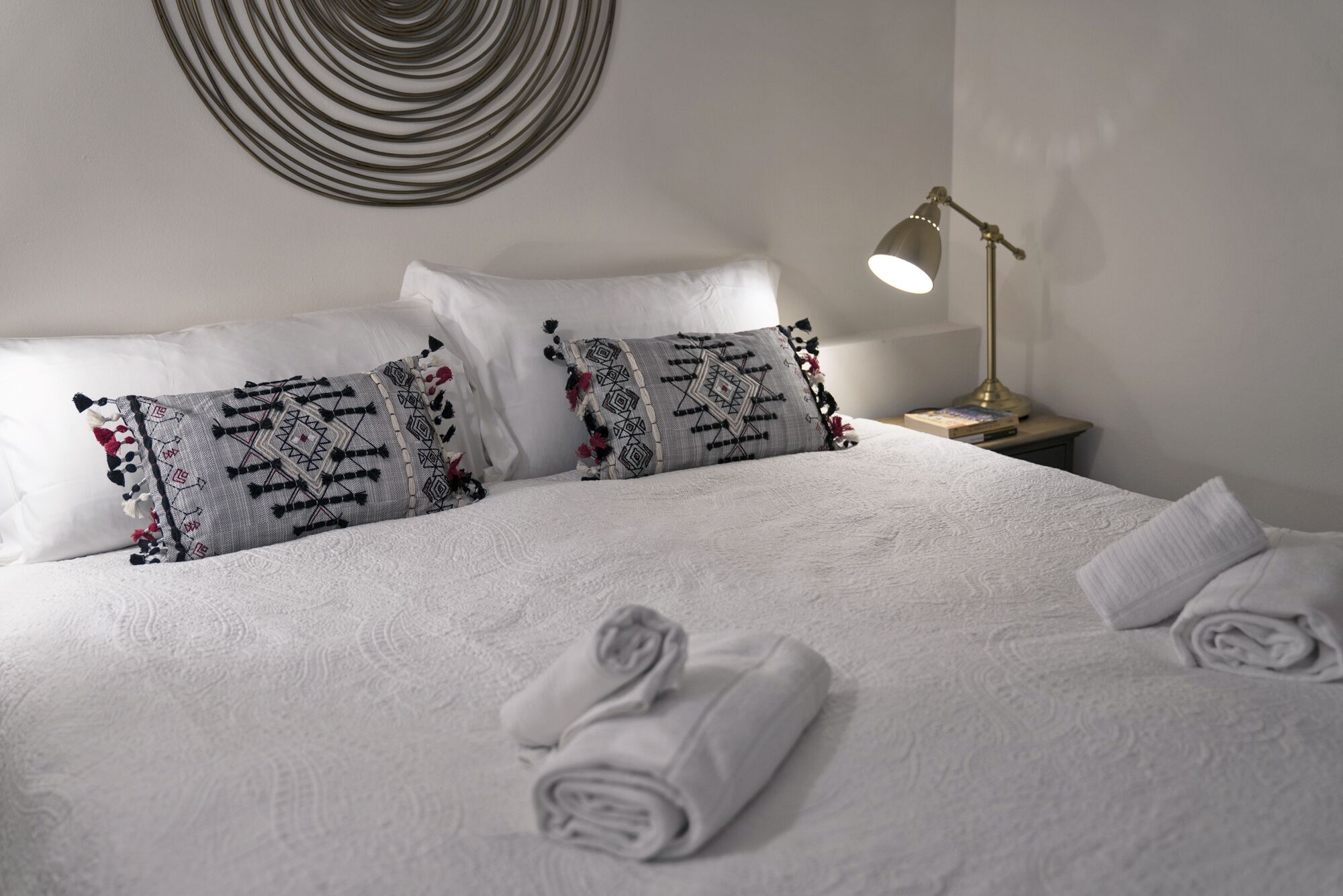 A neatly made bed with patterned pillows, a bedside lamp, and rolled towels on a white textured bedspread.