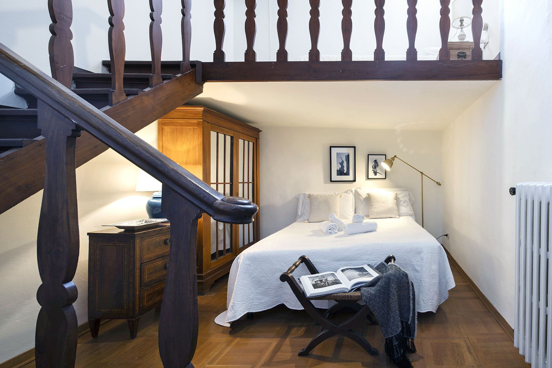 A bedroom with a wooden staircase, a bed with white linens, a chair with a book, and a radiator.