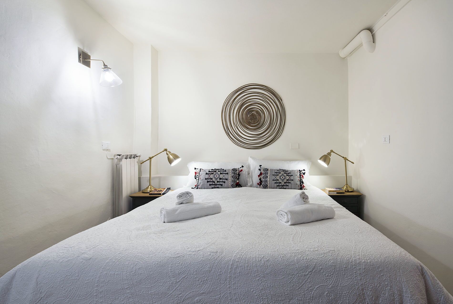 A neatly arranged bedroom with a large bed, two bedside lamps, a wall-mounted circular light, and a radiator.