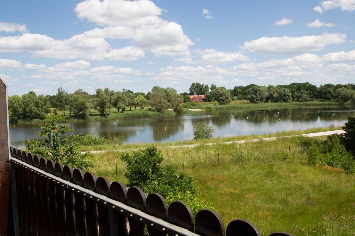 Vista de un lago con una valla de madera en primer plano, árboles y un edificio a lo lejos.