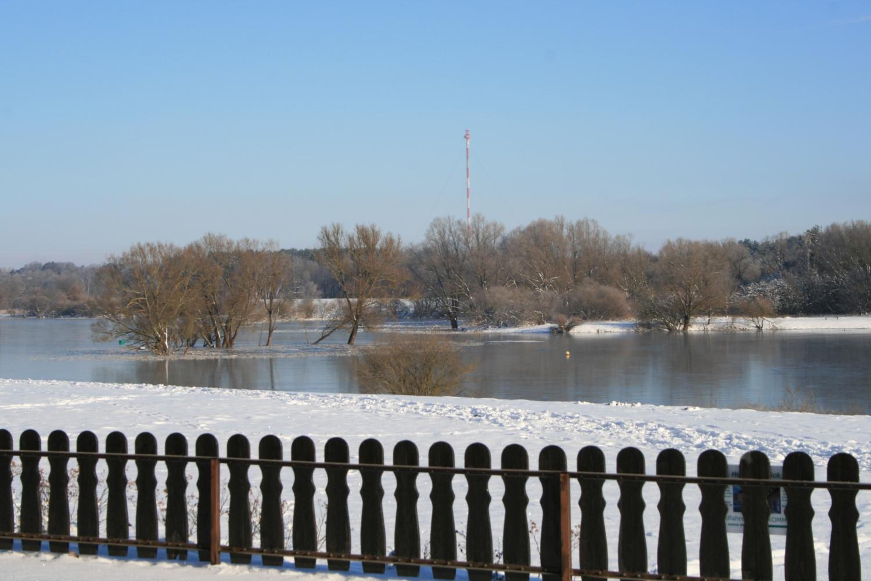 A snow-covered landscape with a body of water, trees, and a fence.