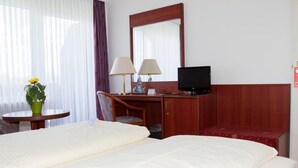 A hotel room with two beds, a desk with a chair, a television, and a small table with a lamp and a flower vase. - Hotel Seeblick Garni (Gartow)