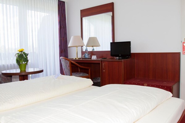 A hotel room with two beds, a desk with a chair, a television, and a small table with a lamp and a flower vase. - Hotel Seeblick Garni (Gartow)