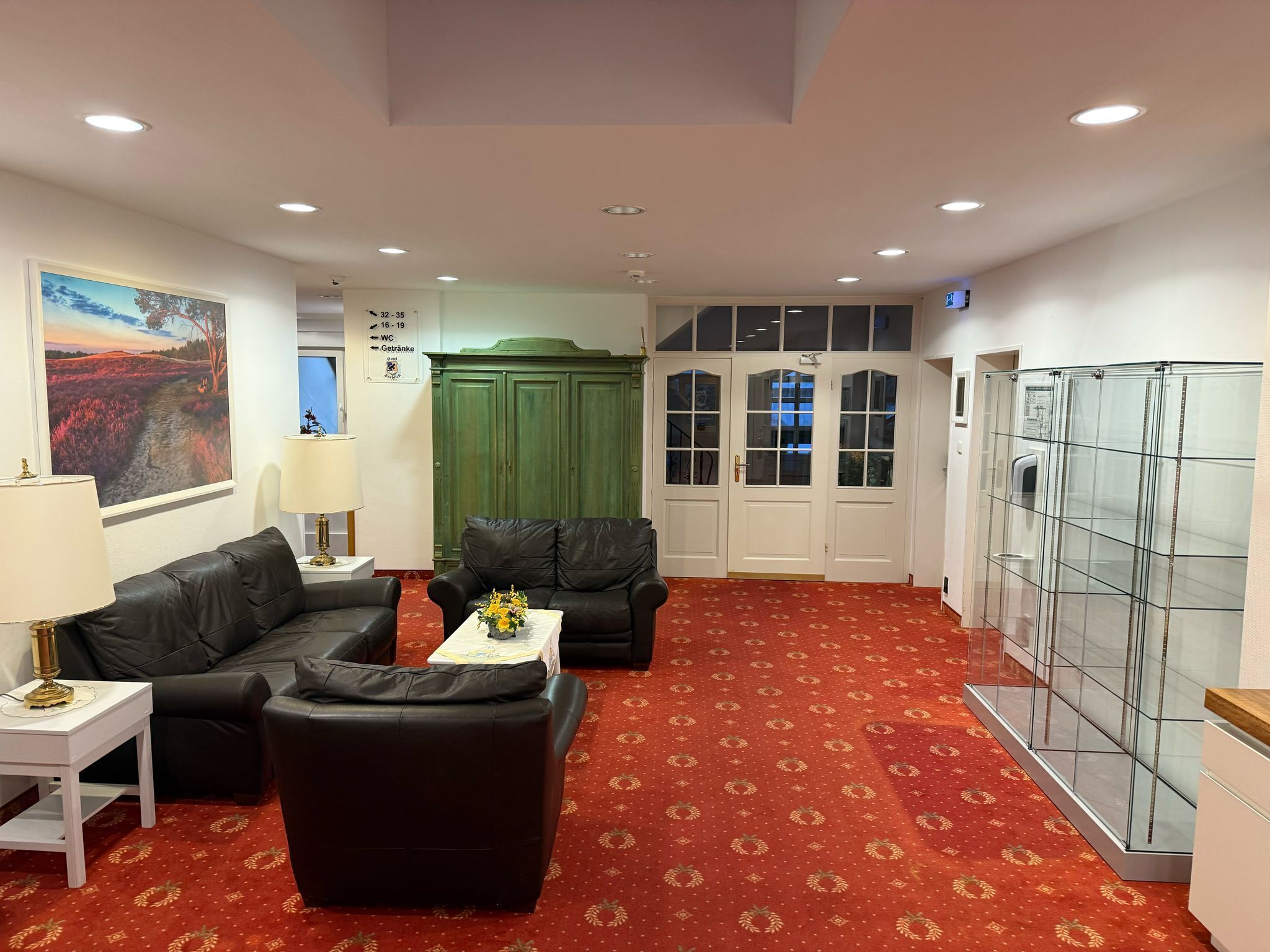 A hotel room with a red carpet, two black leather sofas, a small table with a vase, a green cabinet, and a glass partition.