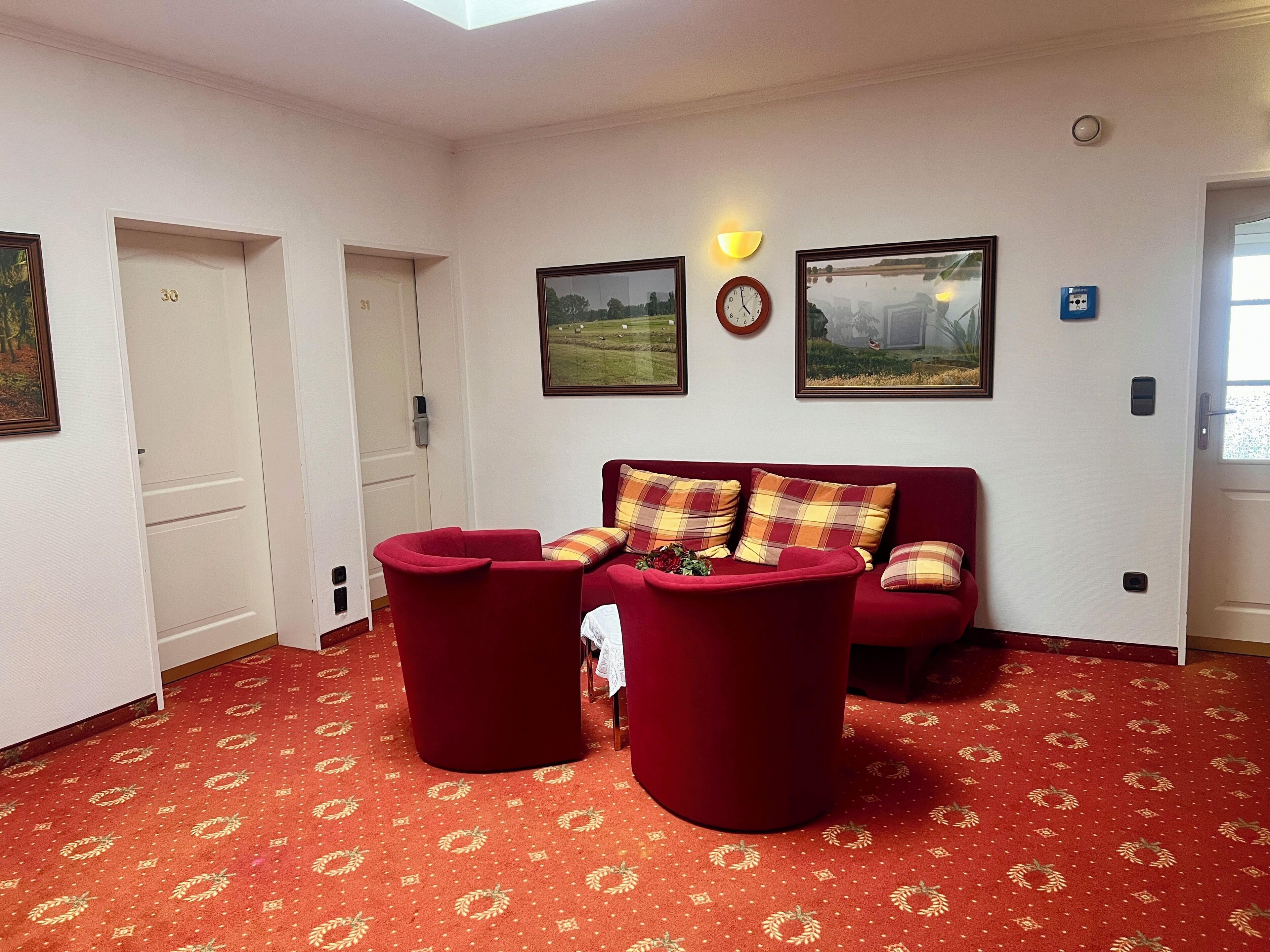 A room with a red carpet, a red sofa, two red armchairs, a wall clock, and two framed pictures.