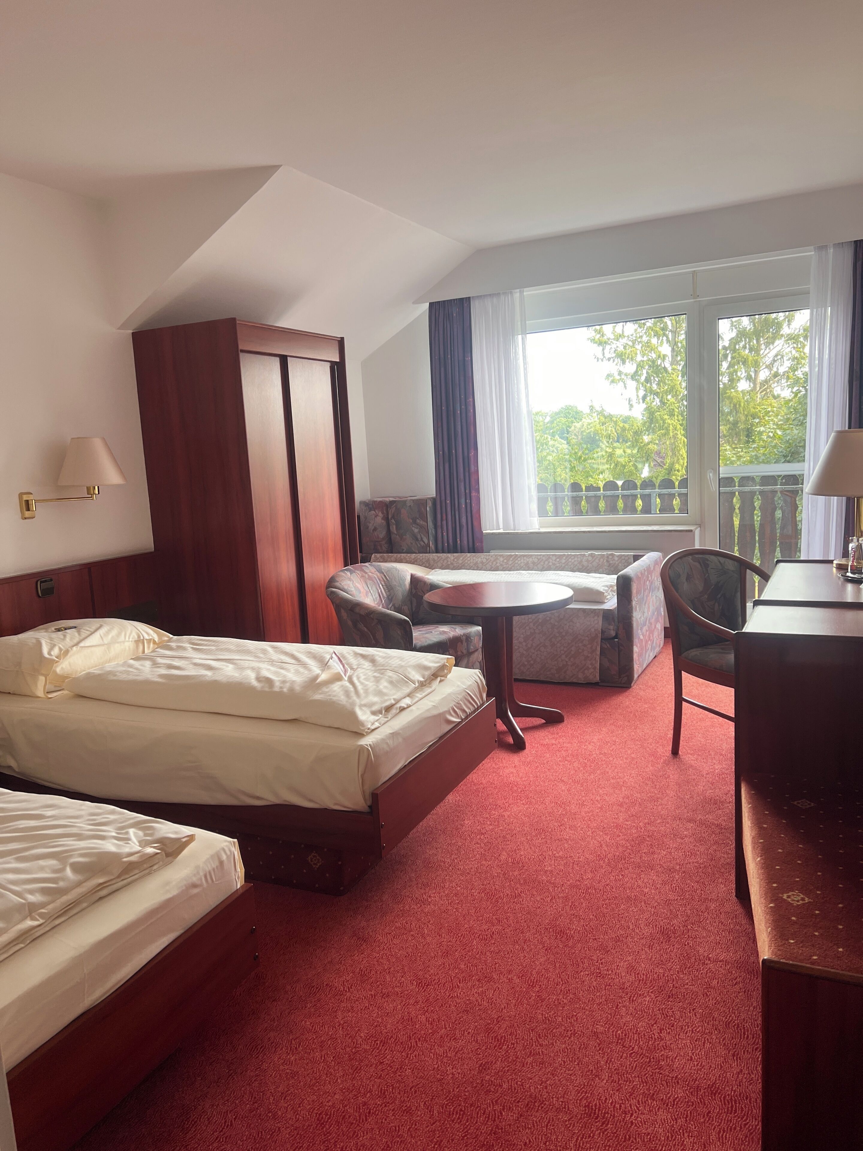 A hotel room with two beds, a desk, a chair, a small table, and a balcony with a view of trees.