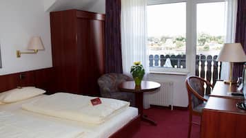 A hotel room with a bed, a desk, a chair, a small table with a plant, and a view of the outside.