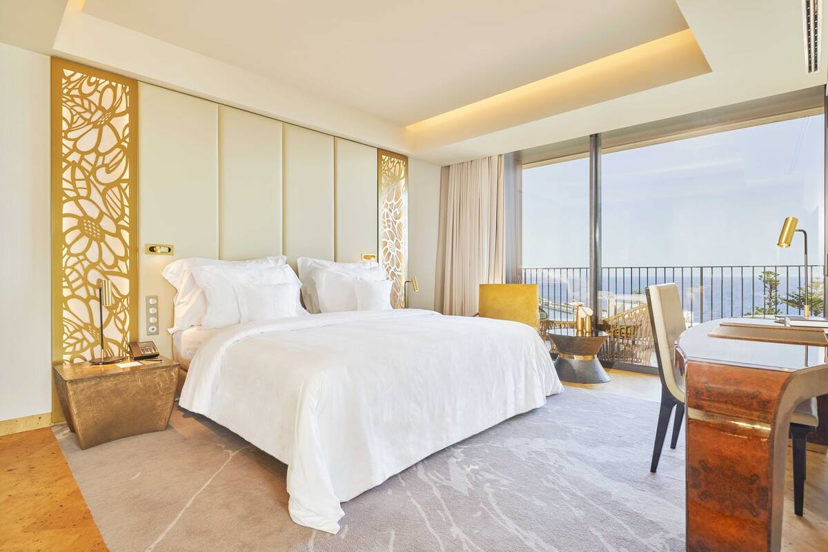A hotel room with a large bed, a desk with a chair, a view of the sea, and a decorative wall panel.