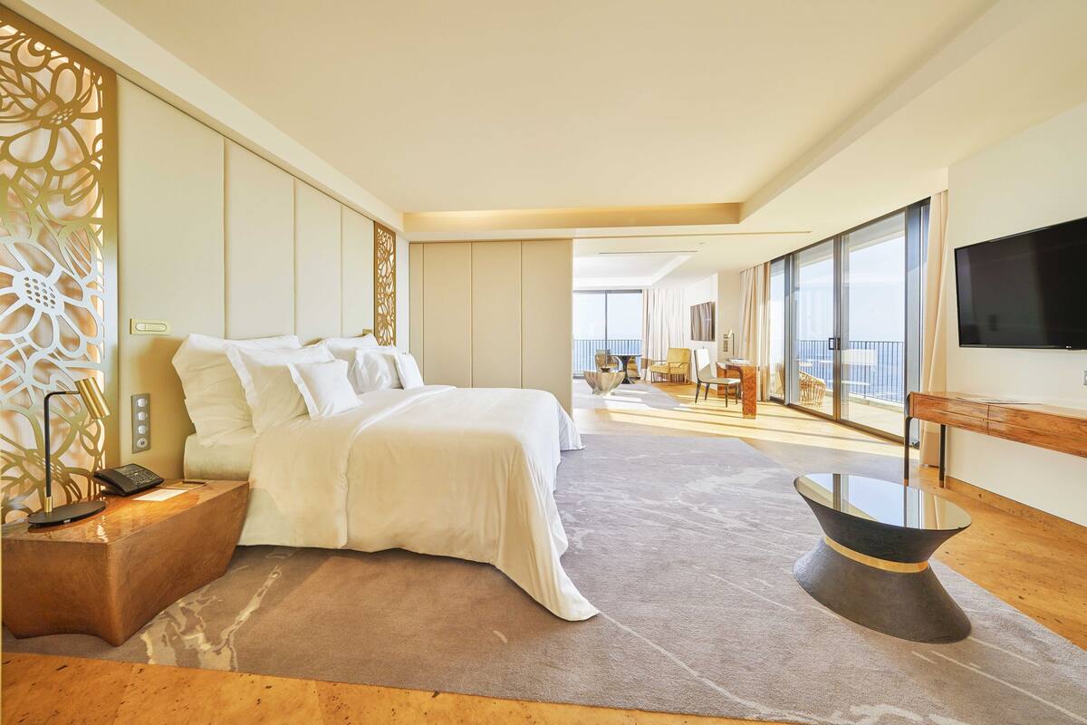 A modern hotel room with a large bed, a bedside table with a phone, a glass coffee table, and a TV on the wall.