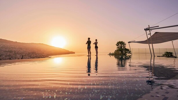 Una pareja de pie en una piscina infinita reflectante al atardecer, con vistas a un paisaje urbano.