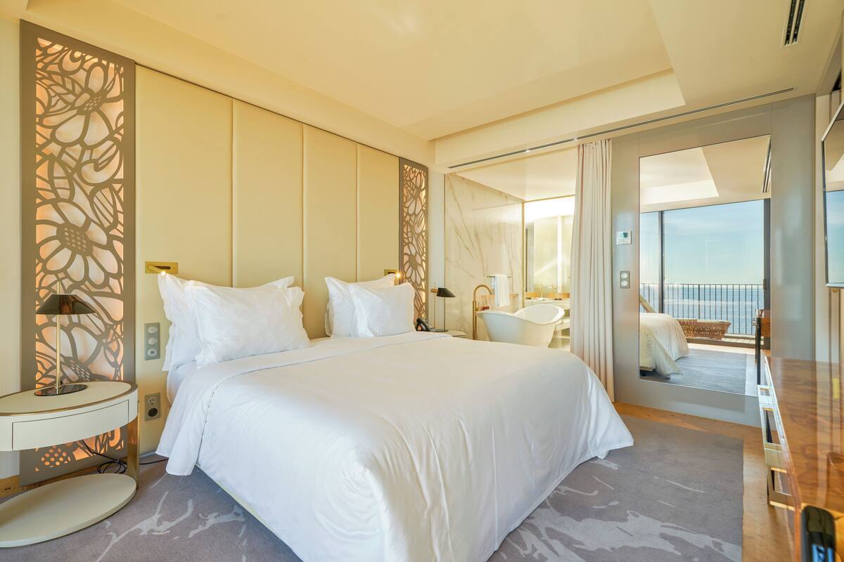 A spacious bedroom with a large bed, a sitting area with a chair and table, and a balcony with a view.