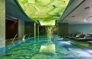 A modern indoor swimming pool with a green ceiling and lounge chairs along the side.