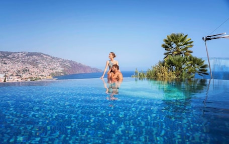 A couple relaxing in a clear infinity pool with a view of a coastal town and hills.