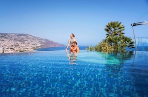 A couple relaxing in a clear infinity pool with a view of a coastal town and hills.