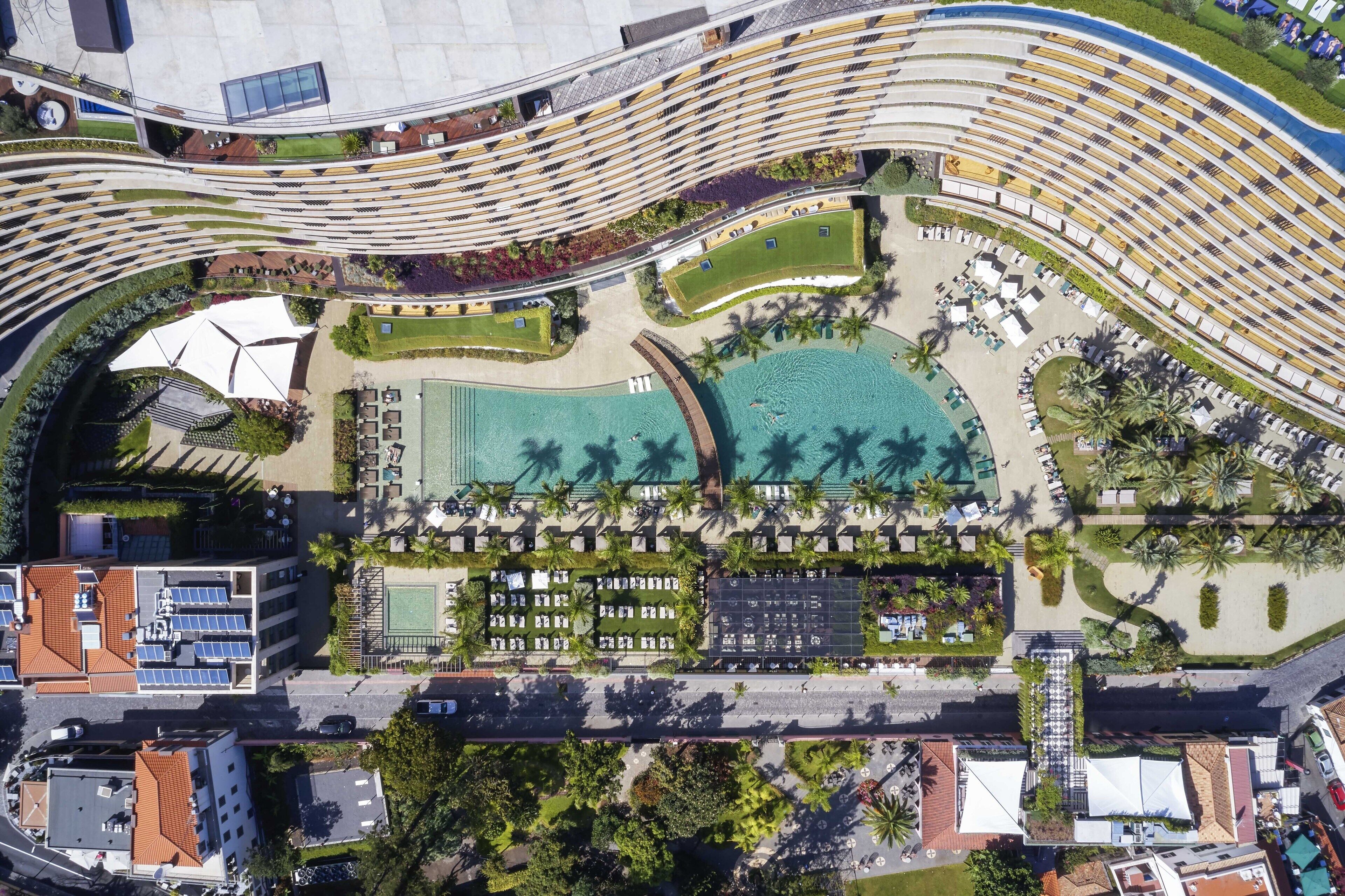 Ein Luftbild eines Hotels mit einem gewundenen Swimmingpool, mehreren Liegestühlen und einem Tennisplatz.