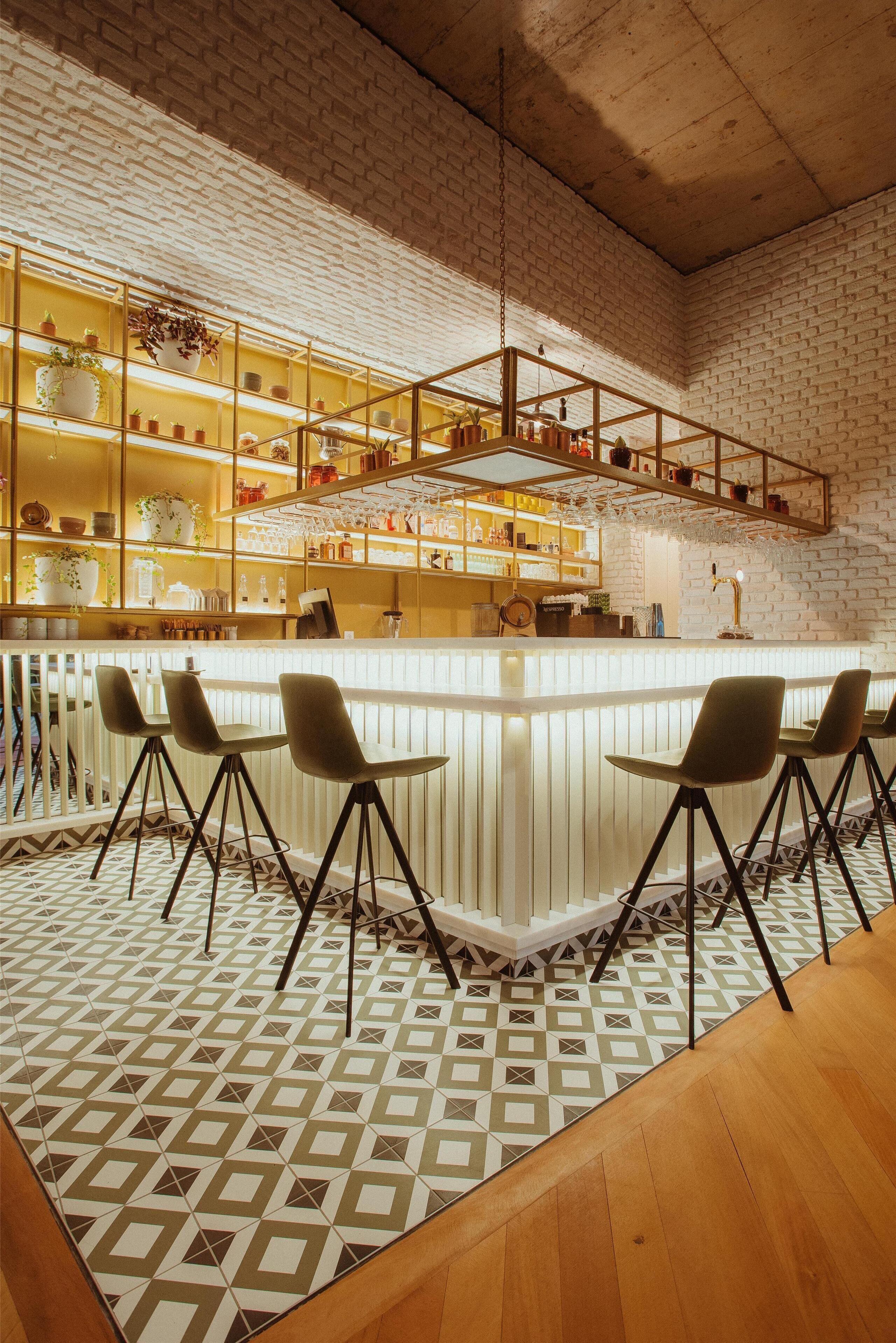 Uma área de bar com balcão branco, vários bancos altos e prateleiras com diversos objetos.