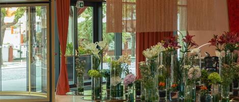 A lobby with large windows, a red curtain, and a series of vases with flowers on a patterned carpet.