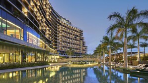 Ein modernes Hotel mit einer geschwungenen Fassade, einem spiegelnden Pool und Palmen.