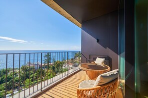 Un balcón con una silla de mimbre y una pequeña mesa, con vistas a la costa, edificios y zonas verdes.