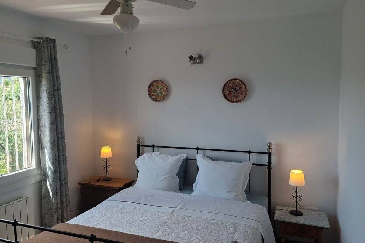 A bedroom with a bed, two bedside tables with lamps, a ceiling fan, and two wall clocks.
