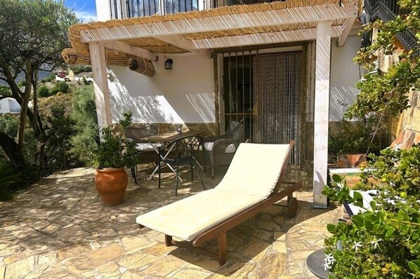 Un espace extérieur ensoleillé, aménagé avec des meubles de jardin, comprenant un fauteuil, une table et des chaises, le tout entouré de plantes en pot et posé sur un sol carrelé en pierre.