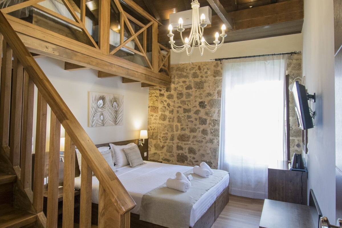 Un dormitorio con escalera de madera, pared de piedra, cama con almohadas, candelabro y televisor.