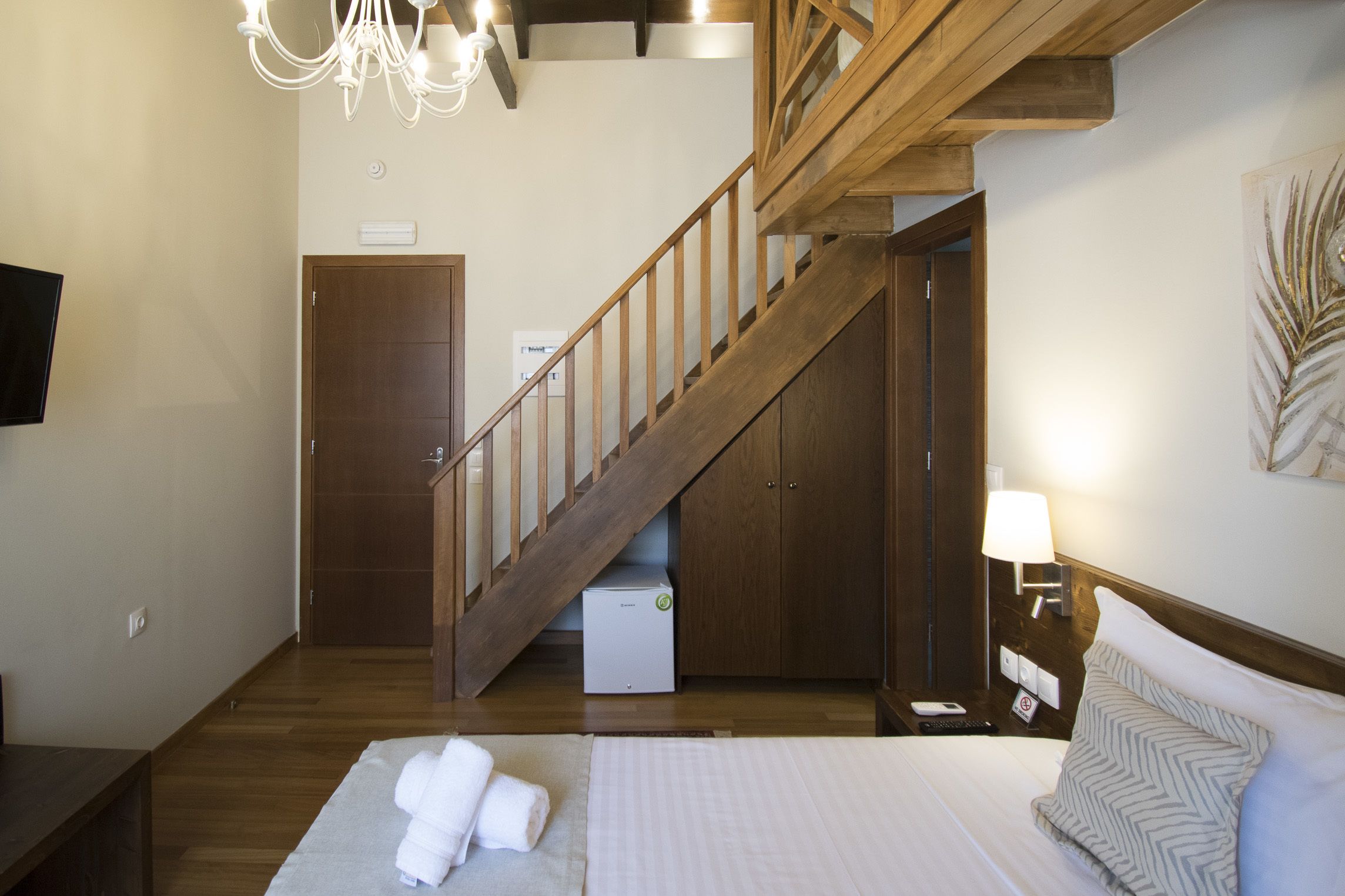 A hotel room with a bed, a wooden staircase, a flat-screen TV, and a wall-mounted lamp.