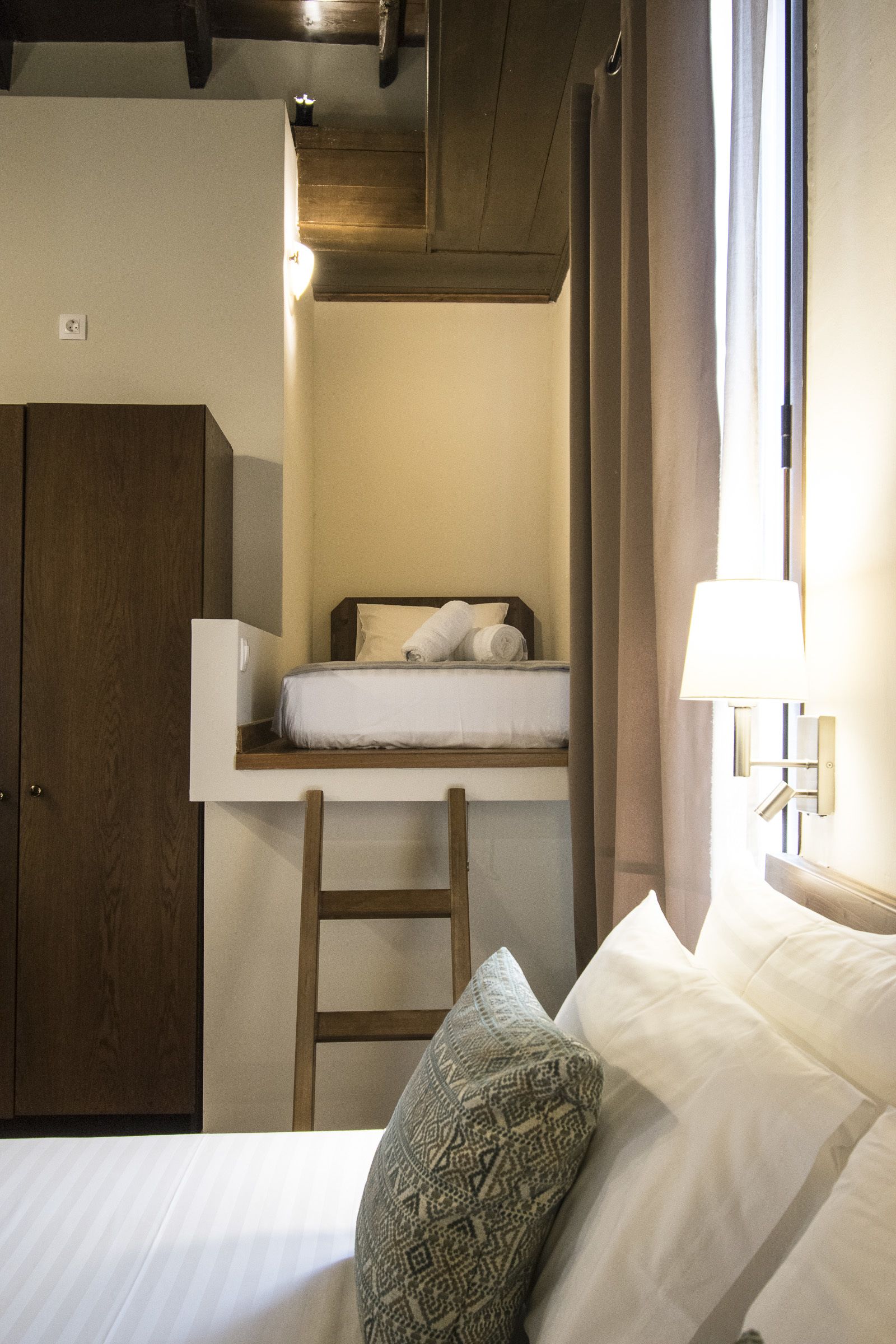 A compact hotel room with a bunk bed, a wooden wardrobe, a bedside table, and a lamp.