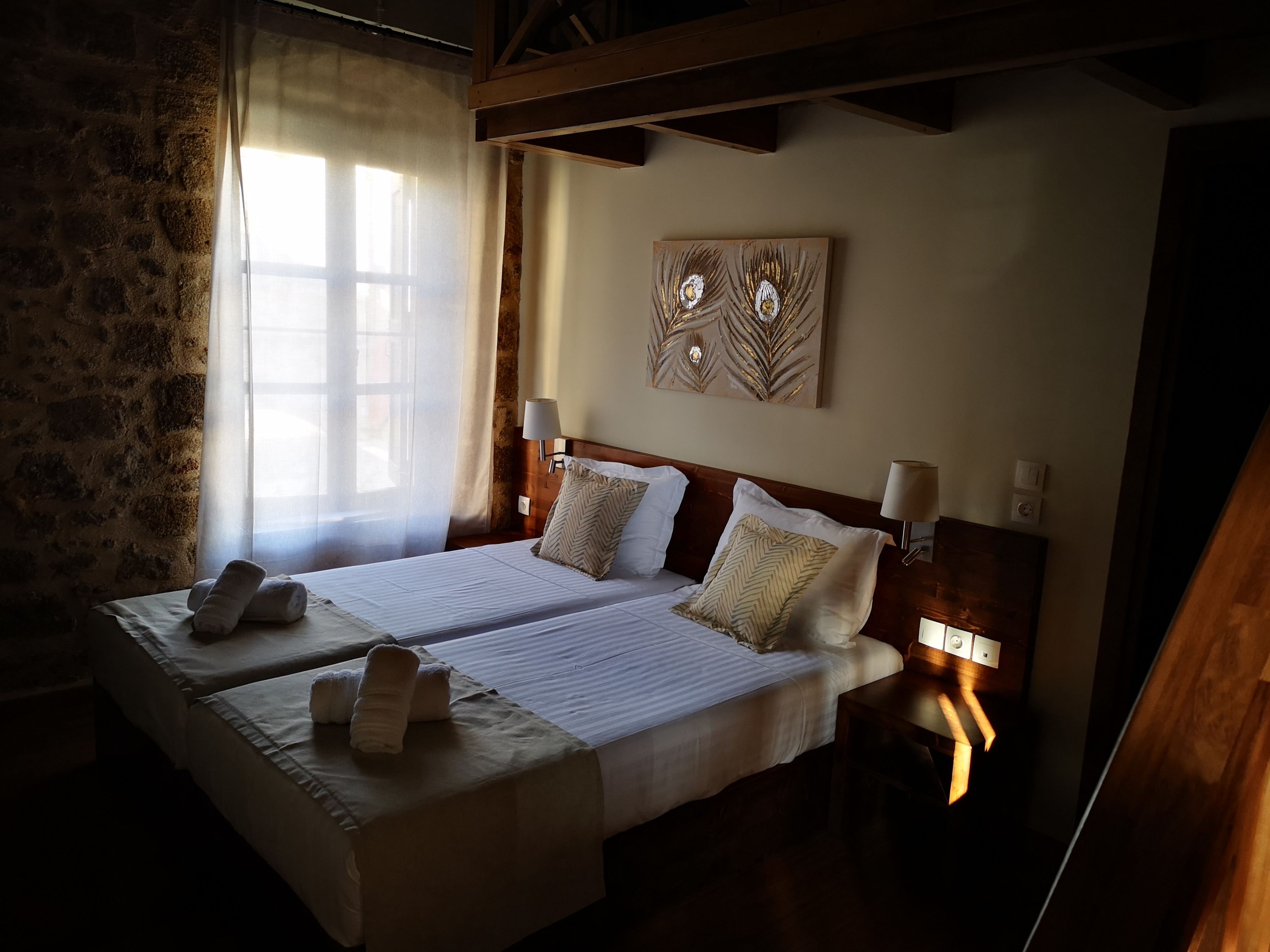 A double bed with two pillows and a blanket, a bedside table with a lamp, a decorative wall piece, and a window with sheer curtains.