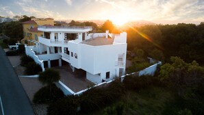 Un edificio moderno de varios pisos con terraza en la azotea, rodeado de abundante vegetación y una vista panorámica al atardecer.