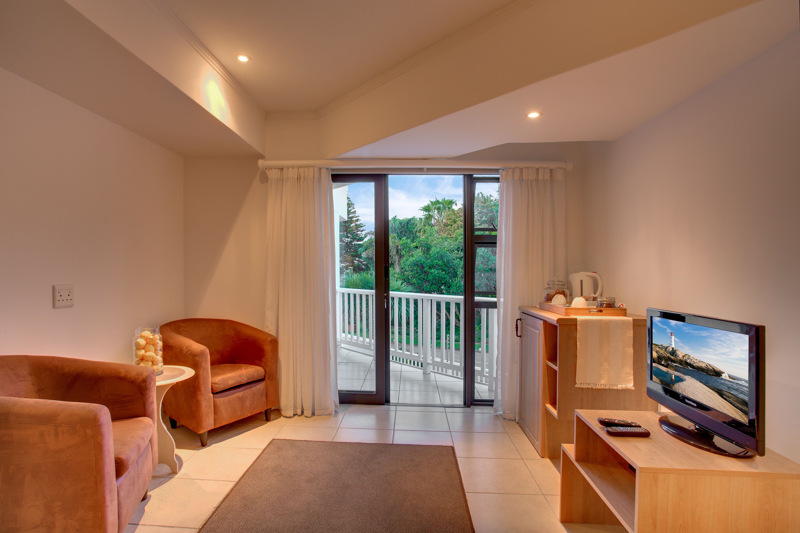 A modern living room with a sofa, a TV, and a balcony with a view of trees and a white fence.