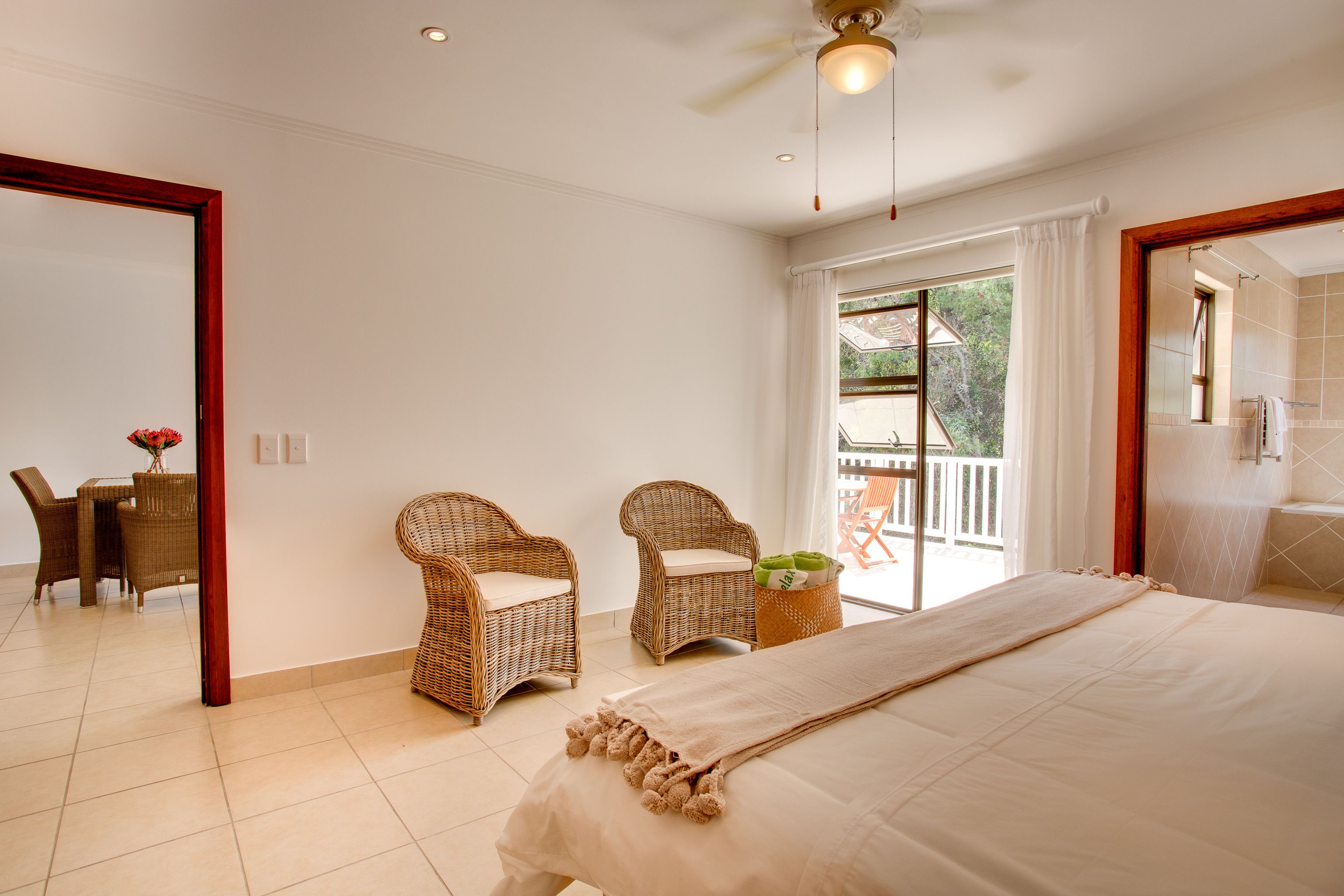 Quarto com cama, duas cadeiras de vime, uma mesinha com um vaso e vista para o exterior.
