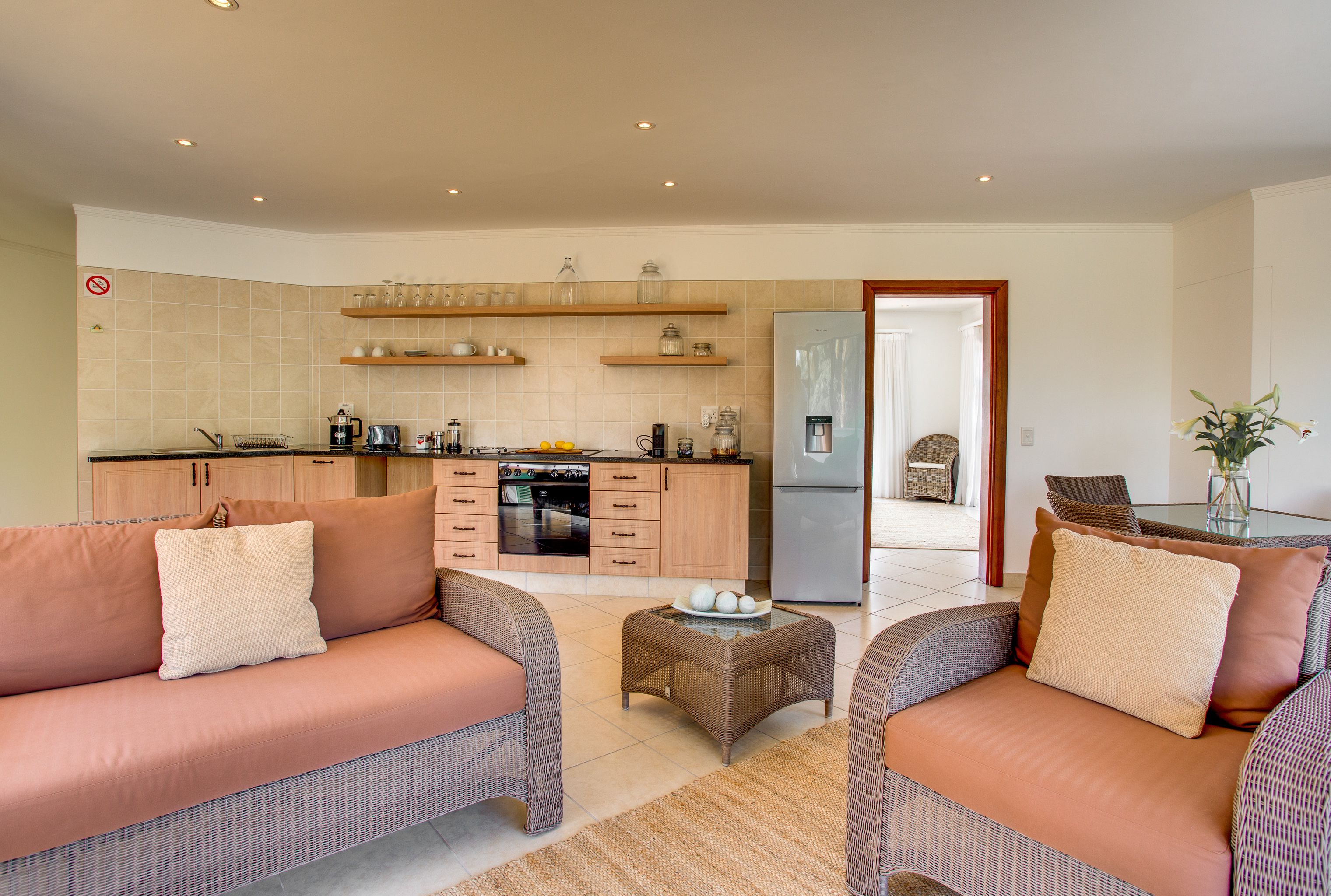 Uma sala de estar moderna com sofá e apoio para os pés, uma cozinha com armários de madeira e geladeira, e uma área de jantar com mesa de vidro.