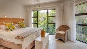 Un dormitorio con una cama grande, una silla de mimbre y vistas a árboles a través de las ventanas.